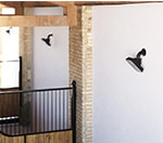 Close-up view of a premium stable interior with Horcery Stall Monitor devices mounted on white walls between brick pillars. The black monitoring units are angled downward toward individual stalls with wooden lower panels and black metal bars.