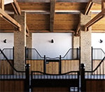A high-end stable interior featuring exposed wooden beams, brick pillars, and modern wood-and-metal stall gates. Horcery Stall Monitor devices are mounted on the walls above each stall, blending advanced technology with traditional barn architecture.