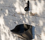 A black Horcery Bucket Meter is mounted on a rustic white wooden wall, suspending a black bucket partially filled with water. The bucket is attached via a metal handle and clip, enabling weight tracking for consumption monitoring in real time.